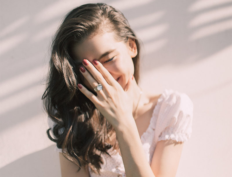 Lachende Frau mit braunen Haaren und weißem Top hält sich die Hand vor den Mund.